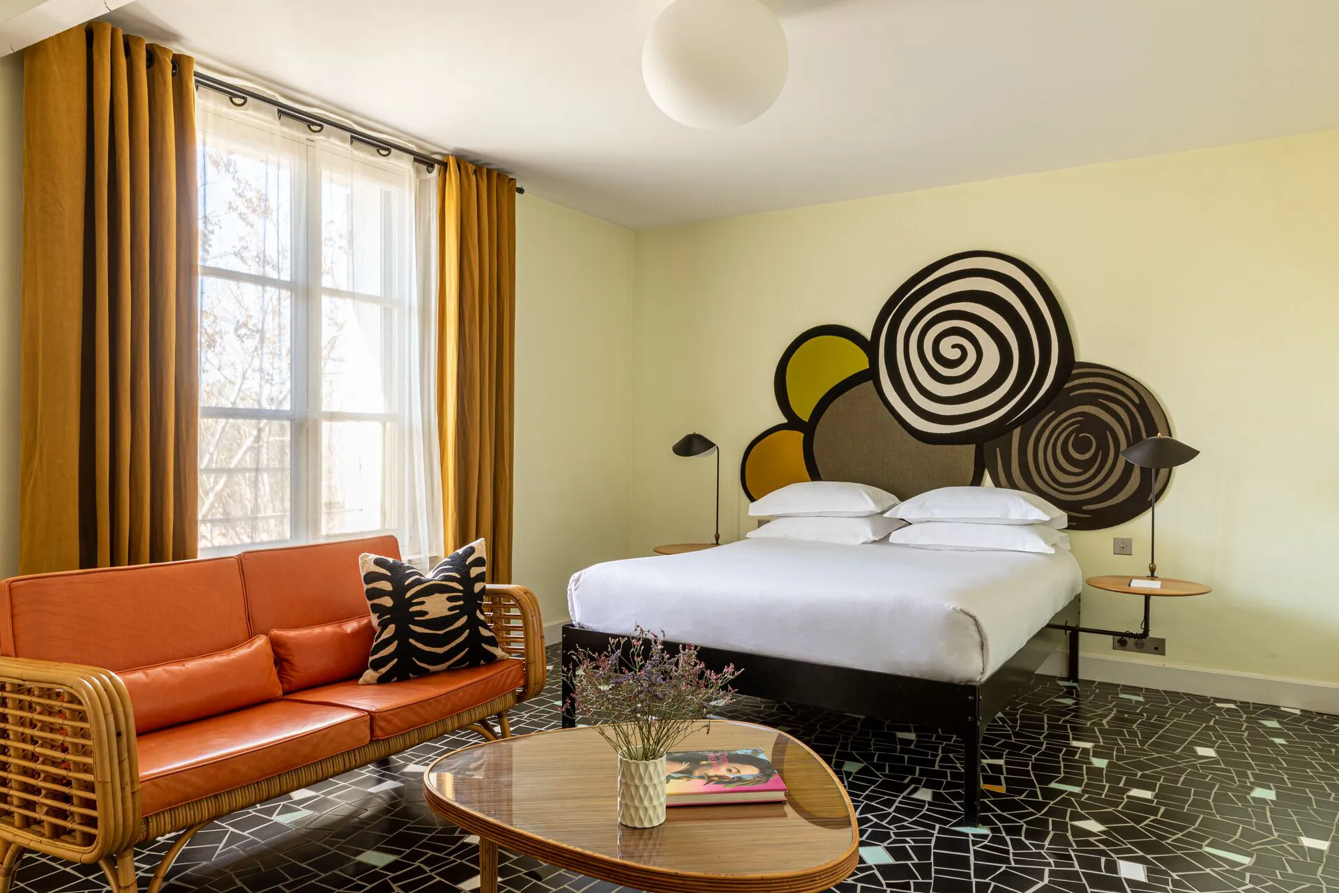 Chambre de l'hôtel Le Cloître. A gauche, une grande fenêtre encadrée de rideaux. Devant la fenêtre un canapé orange en rotin et cuir orange. au centre une table basse et un lit double.