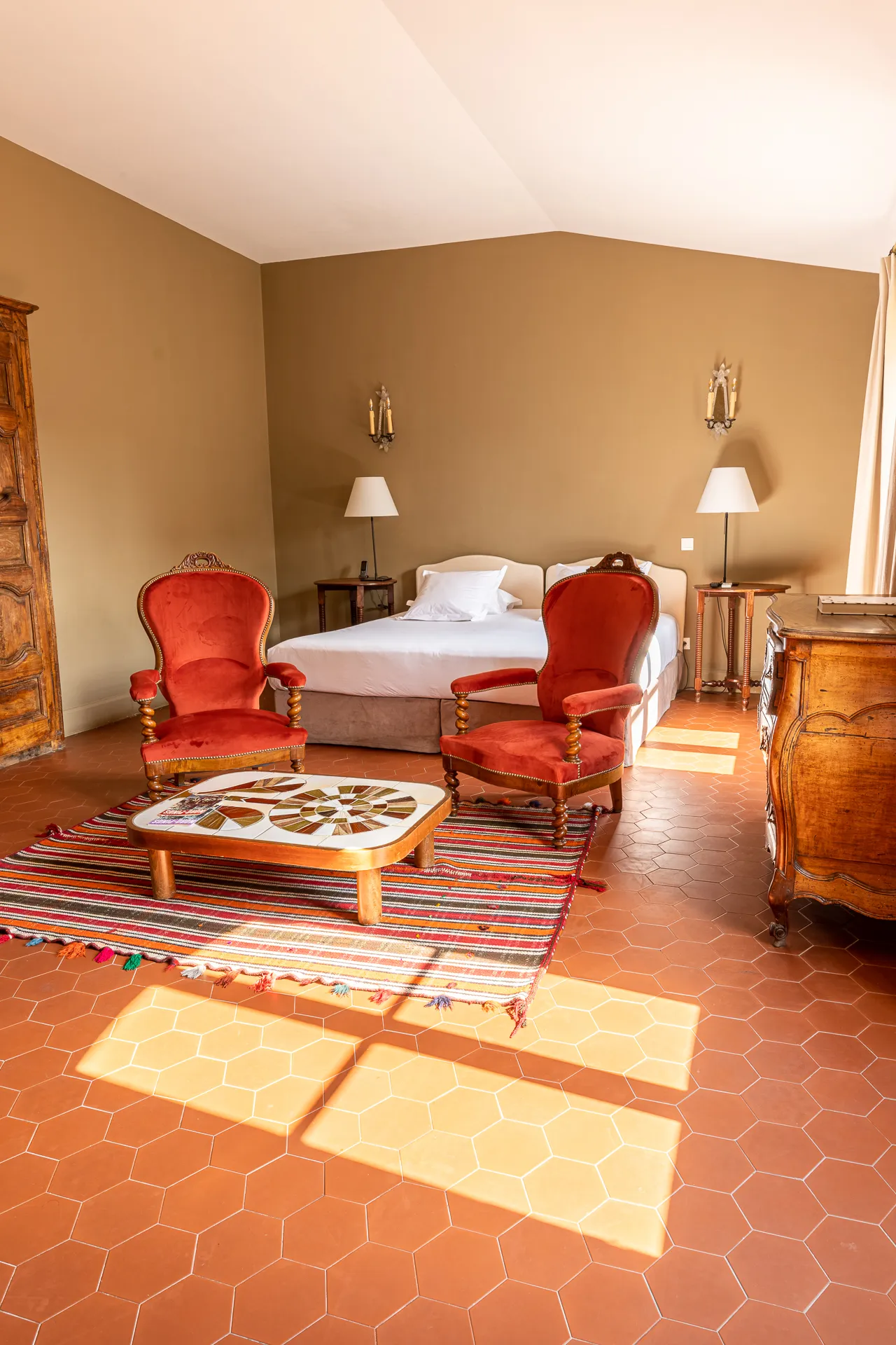 Room at hotel Le Nord-Pinus. On the right, a ray of sunlight streams through the window. The floor is covered with tomettes. In the center of the picture, there are two red Napoleon III armchairs, a coffee table and a bed behind.