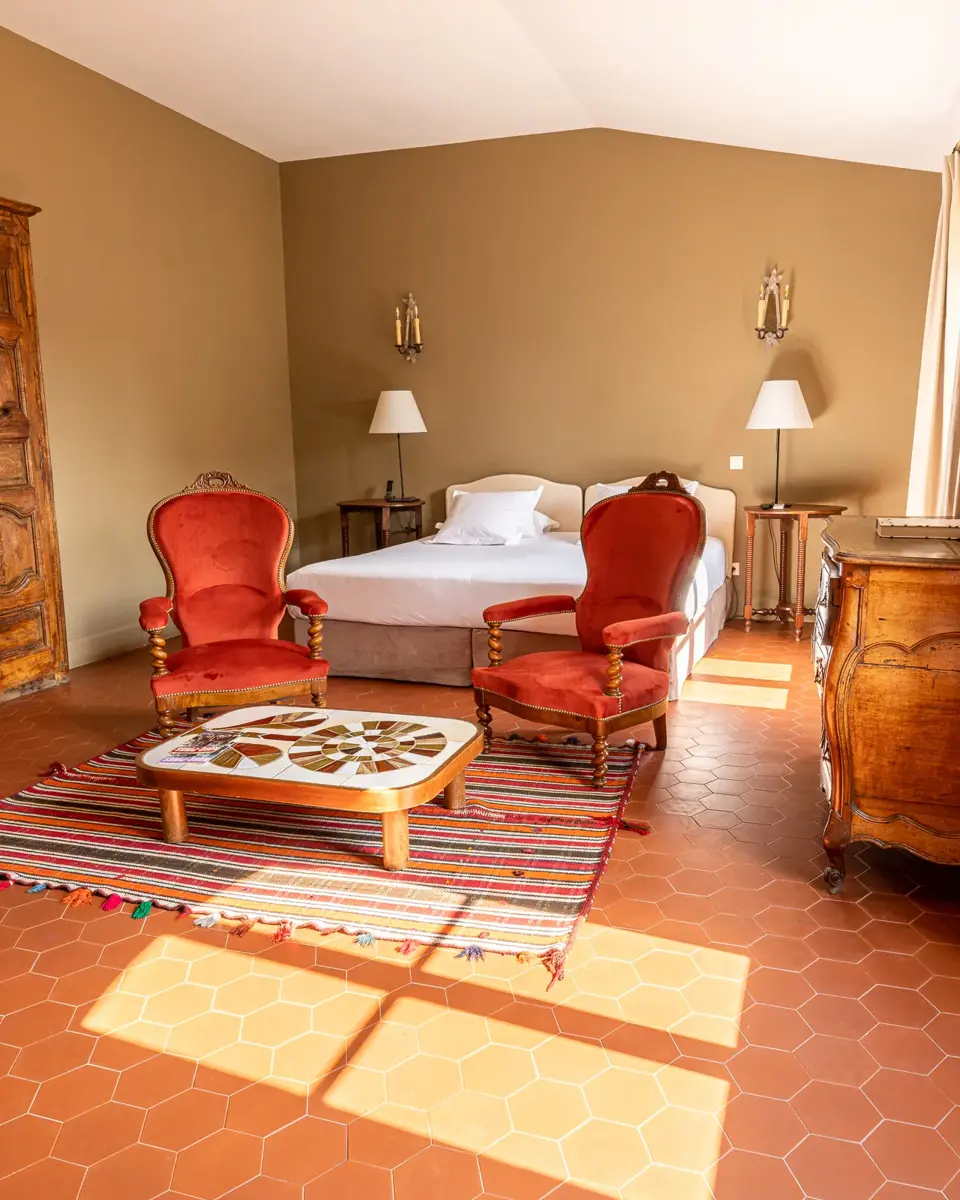 Chambre de l'hôtel Le Nord-Pinus. A droite, un rayon de soleil rentre depuis le fenêtre. Le sol est couvert de tomettes. Au centre de l'image, il y a deux fauteuils Napoléons III rouge, une table-basse et un lit derrière.