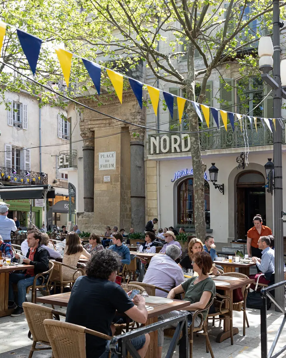 Terrasse du Nord-Pinus sur la place du Forum. Des personnes sont assises autour de tables en bois.