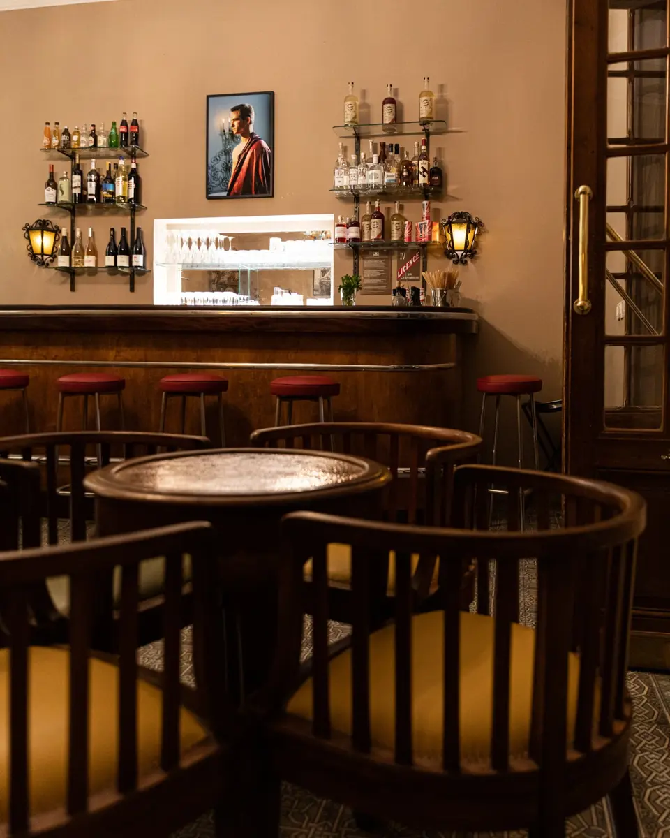 Au premier plan, quatre chaises en bois autour d'une table couverte d'un plateau en cuivre. En arrière plan, un comptoir de bar en bois.