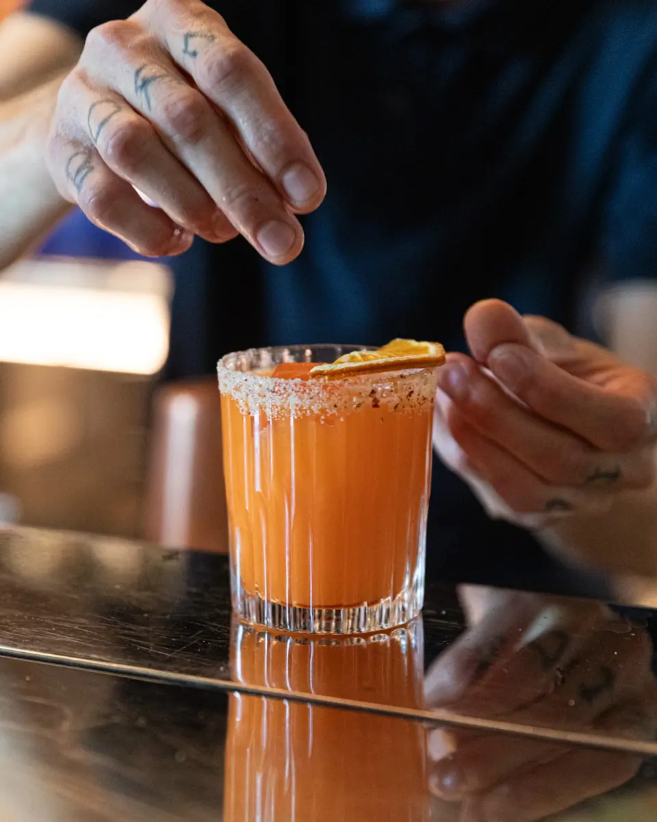 Un cocktail orange posé sur un comptoir en inox. Un barman finalise la préparation.
